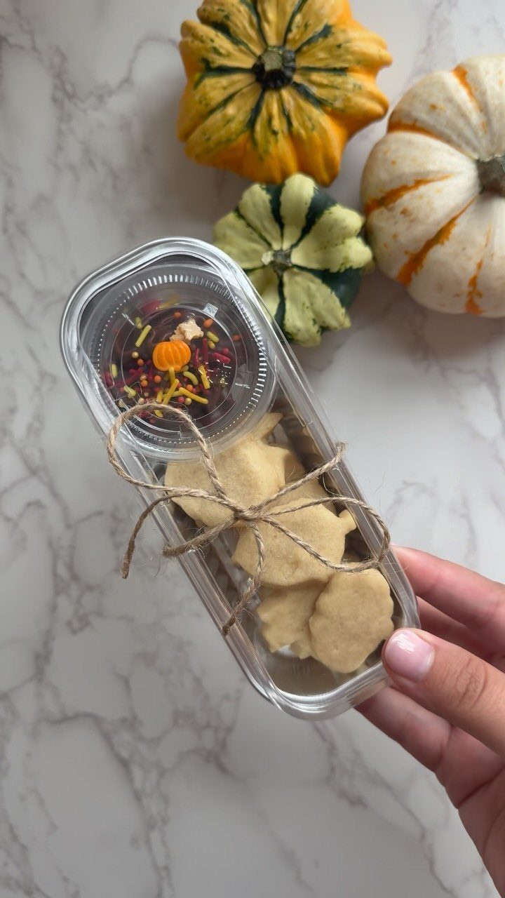 Hand holding a clear container of cookies and sprinkles next to decorative autumn pumpkins.