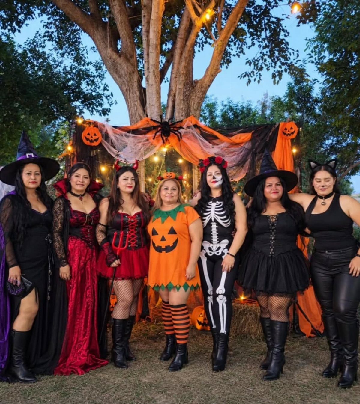 Six women posing in diverse Halloween costumes in front of a decorated tree.