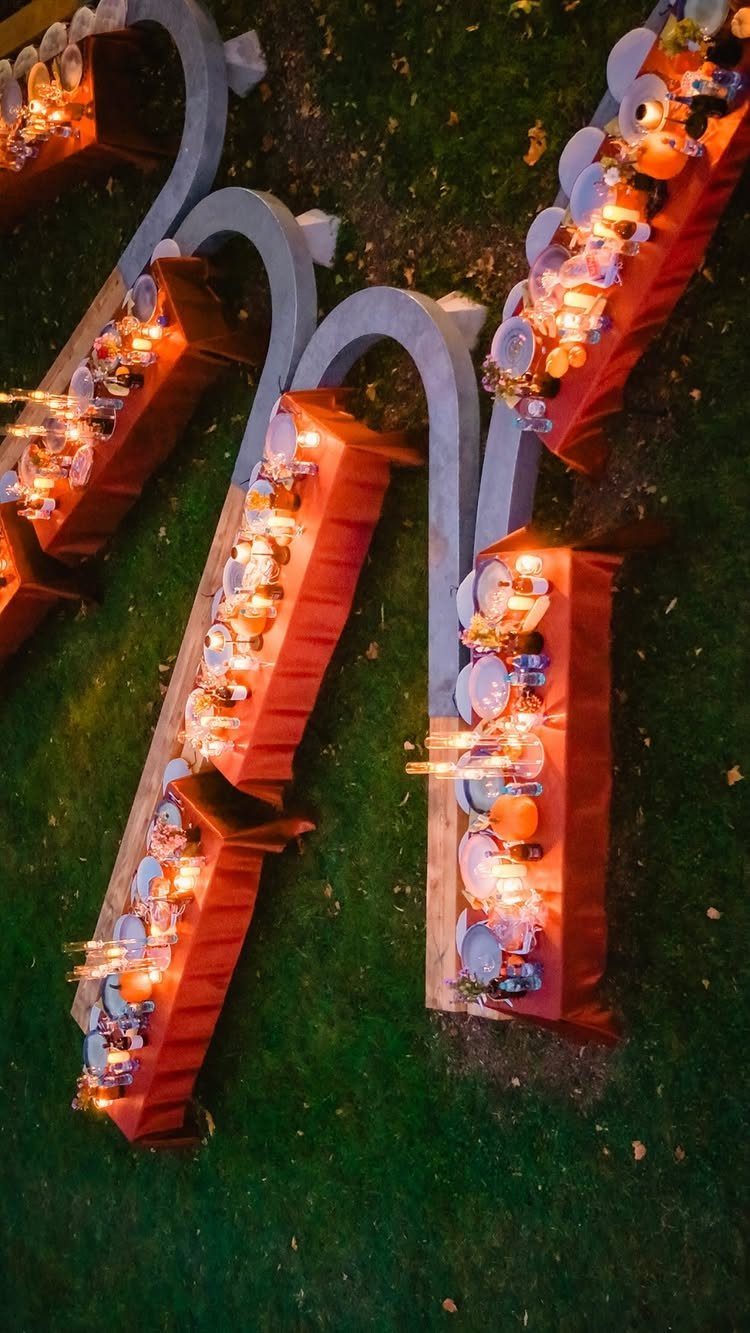 Long outdoor dining tables with orange linens and glowing candles arranged on a green lawn at night.