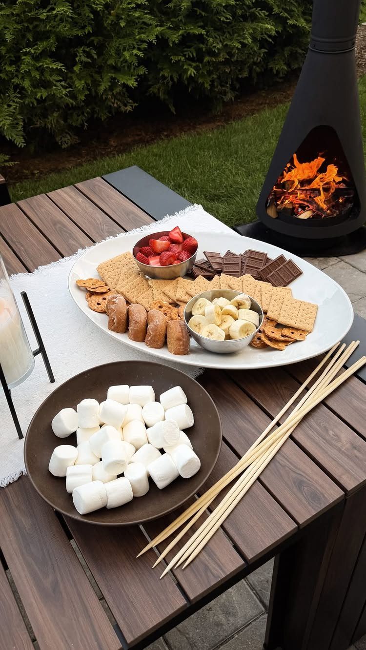 S'mores ingredients including marshmallows, chocolate, graham crackers, and fruit arranged on a table near a bonfire.