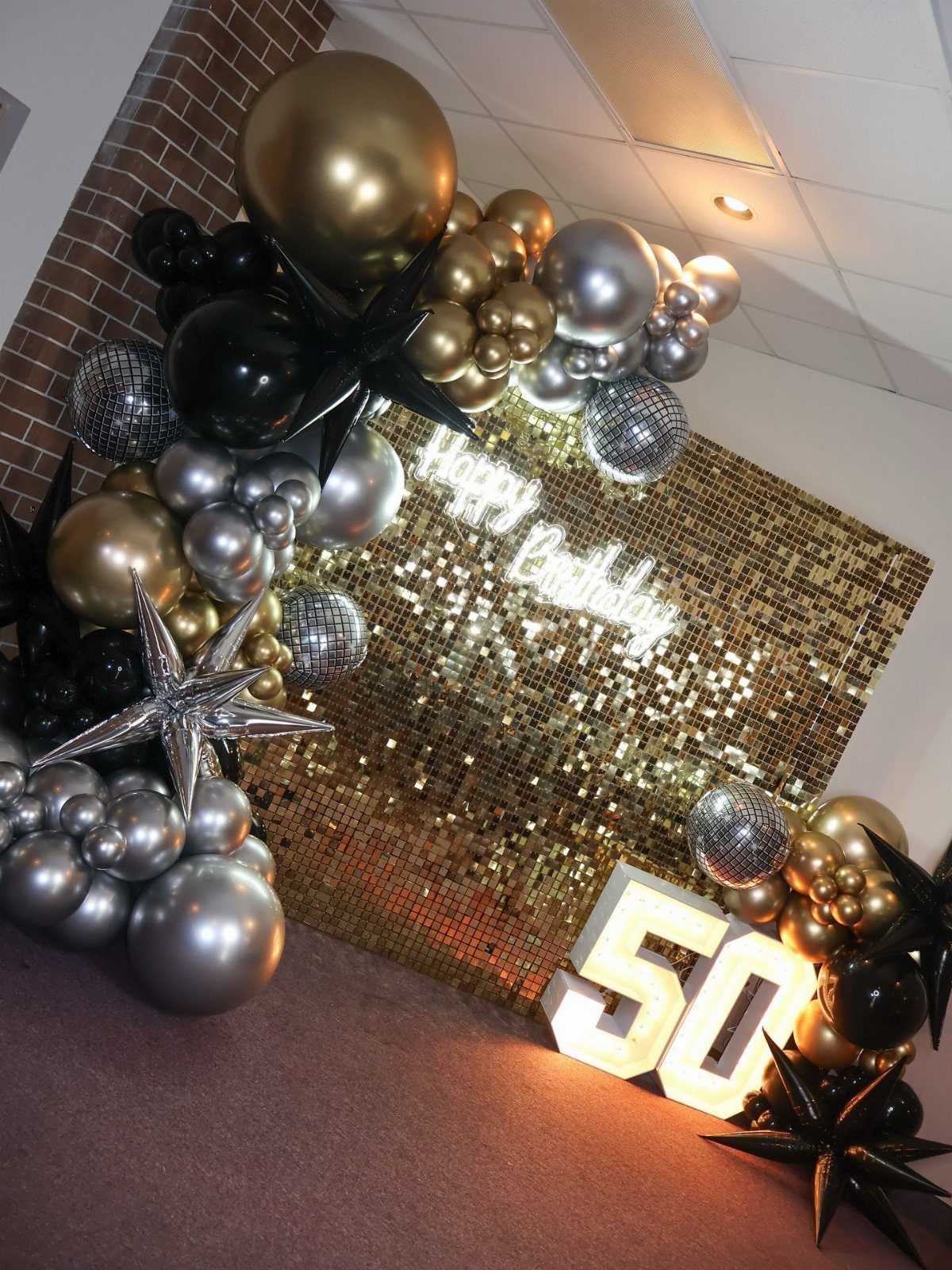 Gold sequin backdrop with black and silver balloons and large illuminated number 50.
