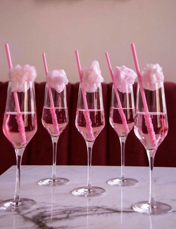 Five champagne flutes with pink liquid, pink straws, and pink cotton candy on marble.