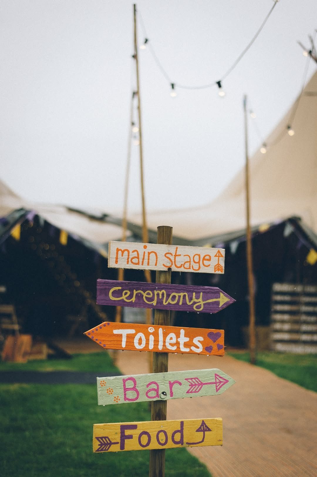 Colorful wooden directional signs pointing toward a main stage, ceremony, bar, and food at an outdoor event.