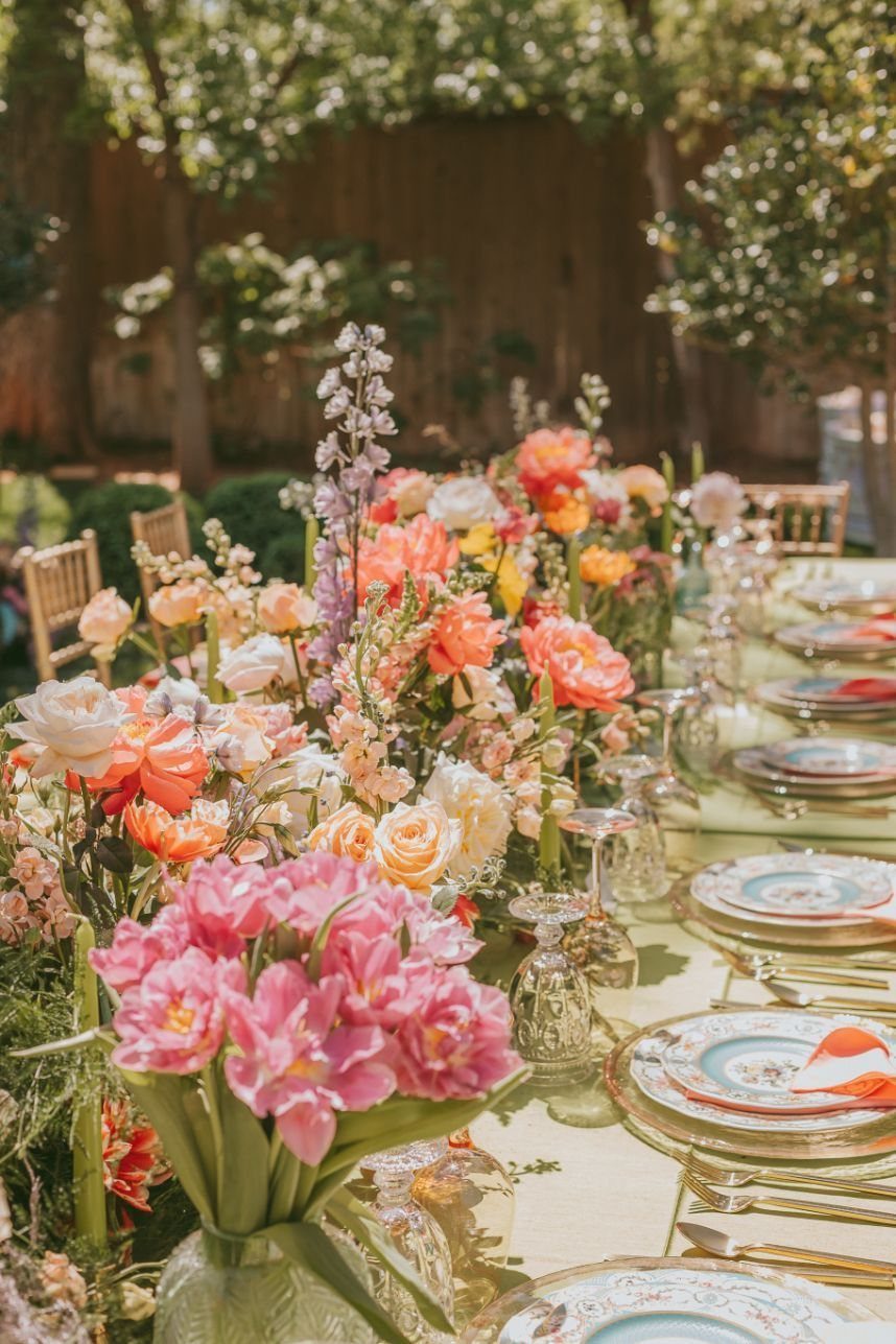 A long dining table decorated with vibrant floral arrangements and elegant place settings in a garden.