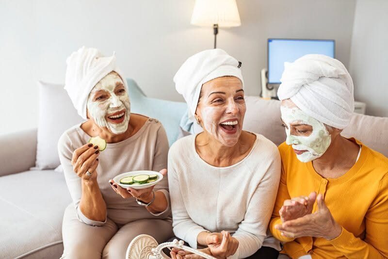 Three women wearing facial masks and hair towels laughing together on a sofa.