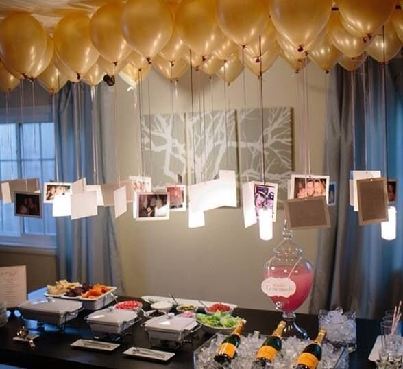 Gold balloons on a ceiling with hanging photographs above a buffet table with food and drinks.
