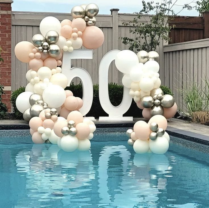 Large white number fifty decorated with cream and gold balloons by a swimming pool.