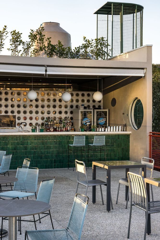 A green tiled rooftop bar with liquor bottles, patio tables, and chairs under a clear sky.