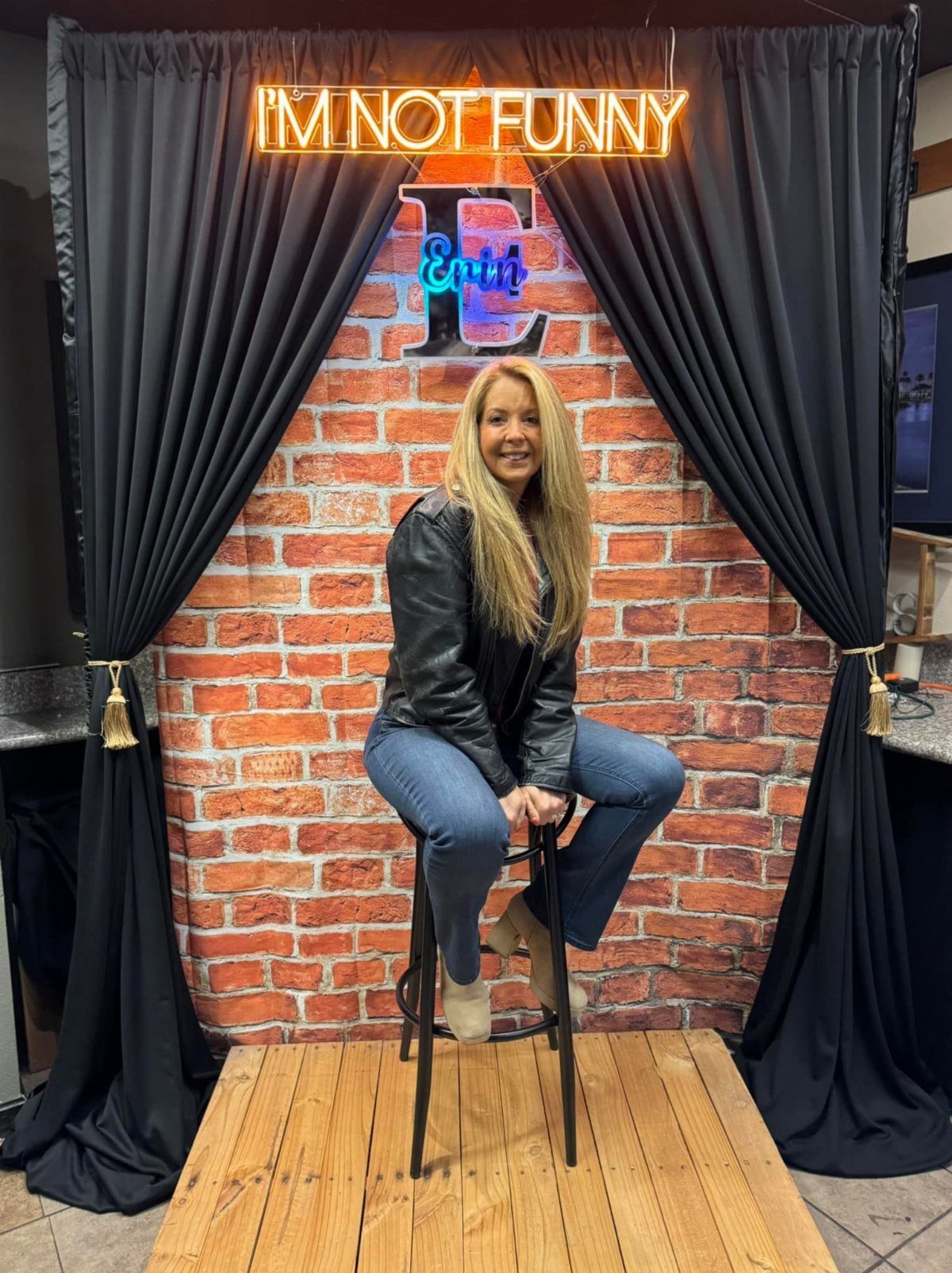 A blonde woman sitting on a stool in front of a comedy club themed backdrop.