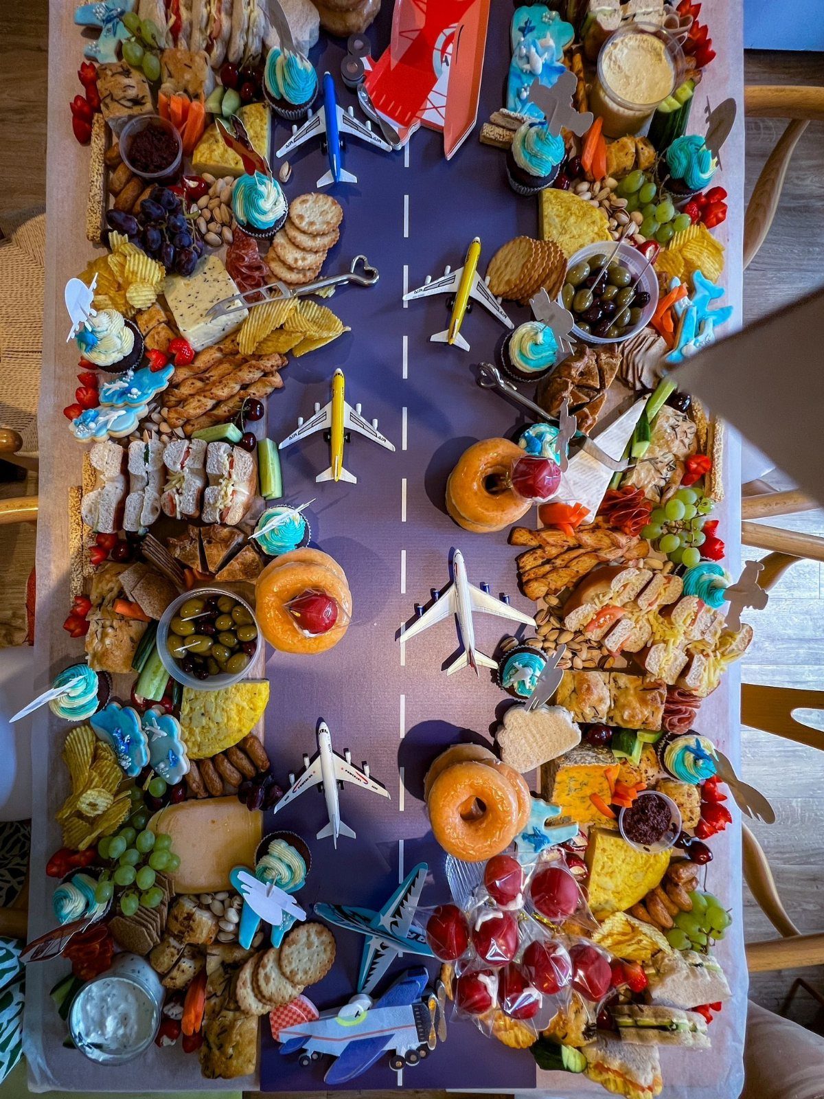 A long grazing table featuring a purple runway with toy airplanes surrounded by assorted snacks and cupcakes.