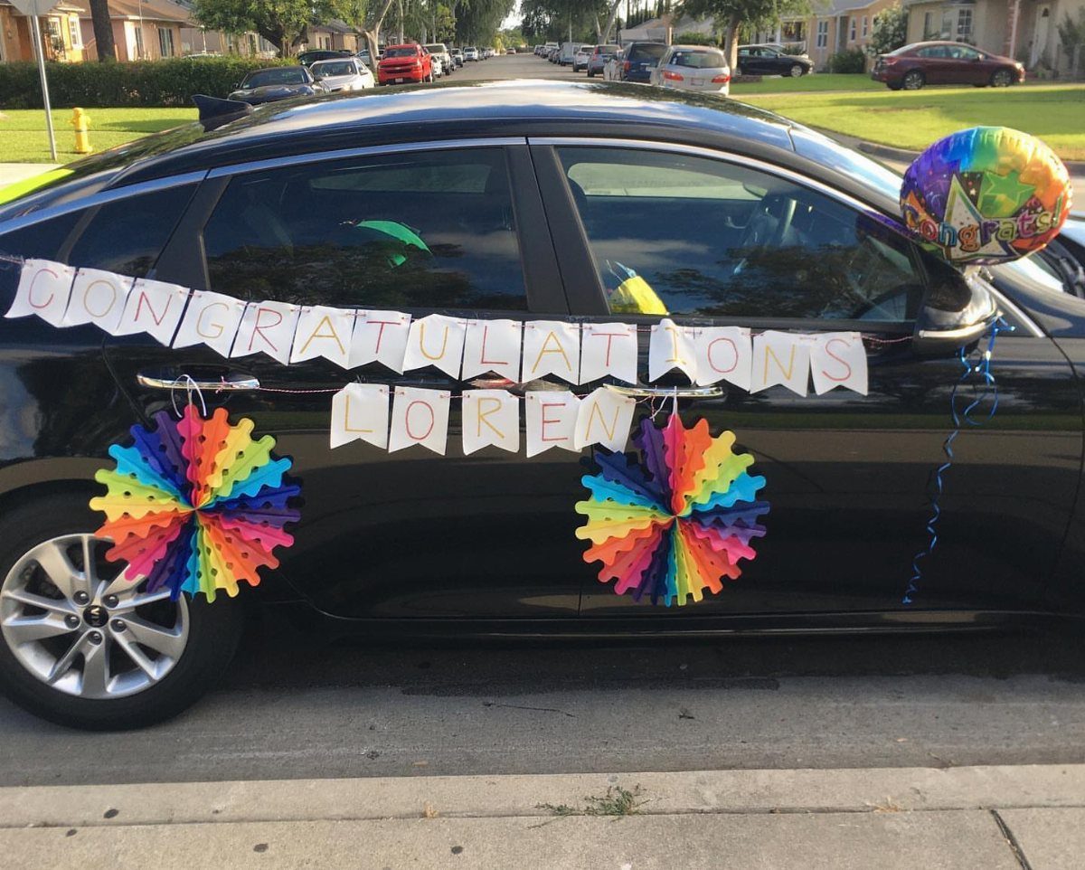 A black car decorated with a congratulations banner, rainbow paper fans, and a colorful balloon.