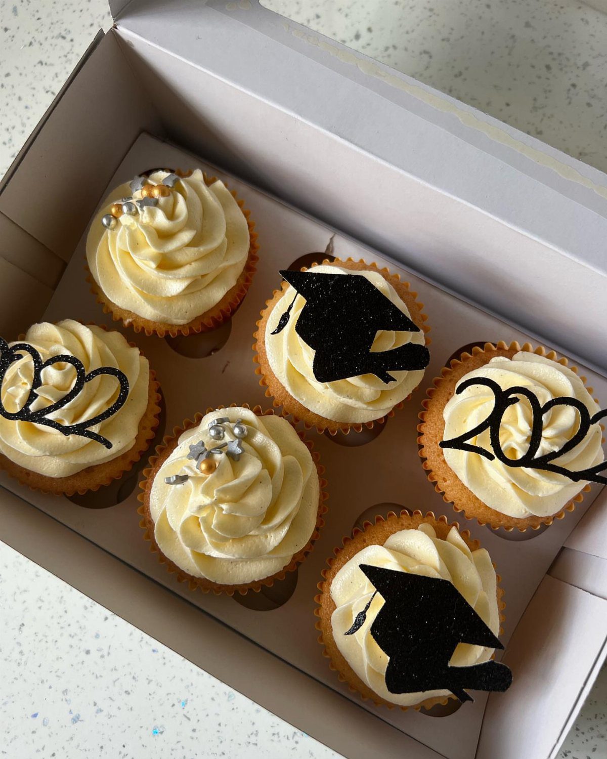Six cupcakes with graduation themed toppers inside a white cardboard box.