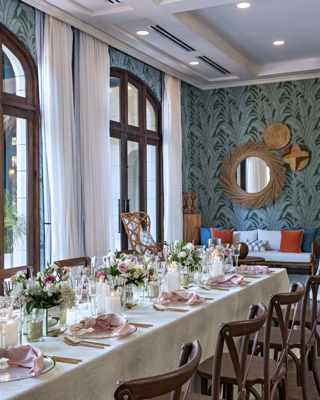 A long dining table set with elegant floral arrangements, pink napkins, and gold cutlery in a tropical room.