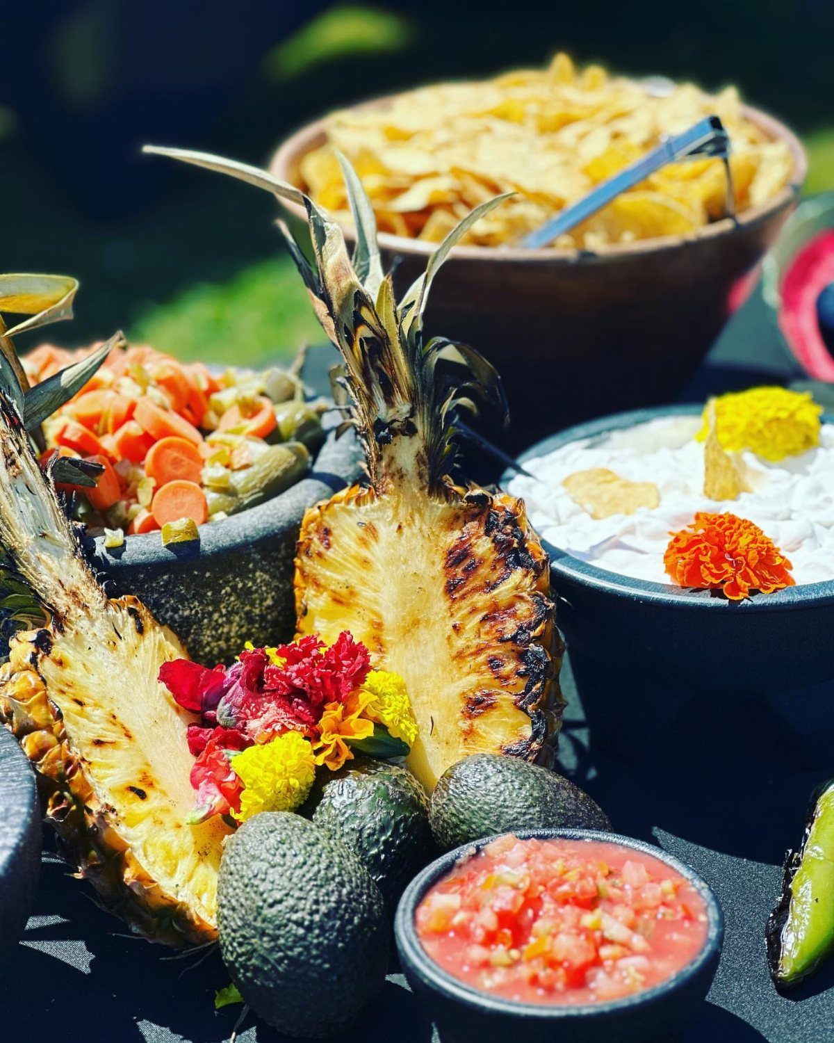 Grilled pineapple, avocados, and salsa bowls arranged on a colorful outdoor food table.