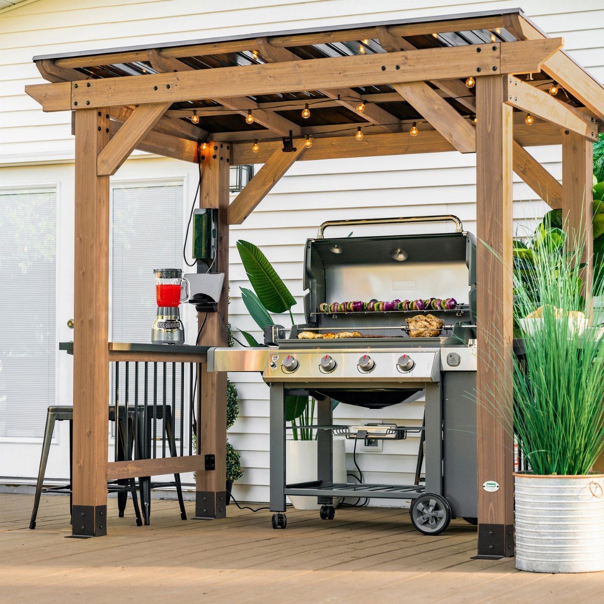 A stainless steel gas grill cooking food under a wooden outdoor kitchen pergola.