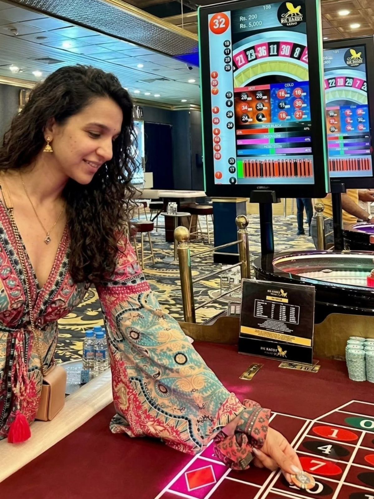 A woman in a patterned dress points at a roulette table in a casino setting.