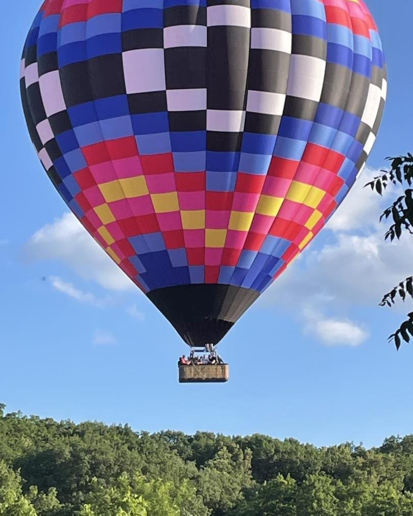 A colorful checkered hot air balloon with a basket of people flying over a green forest.