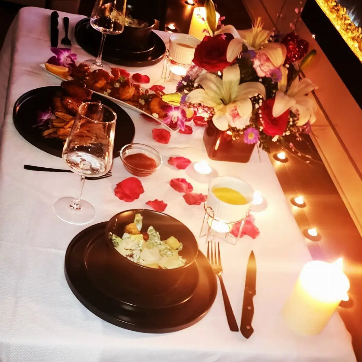 An elegantly set dining table with white lilies, red roses, rose petals, and lit candles.