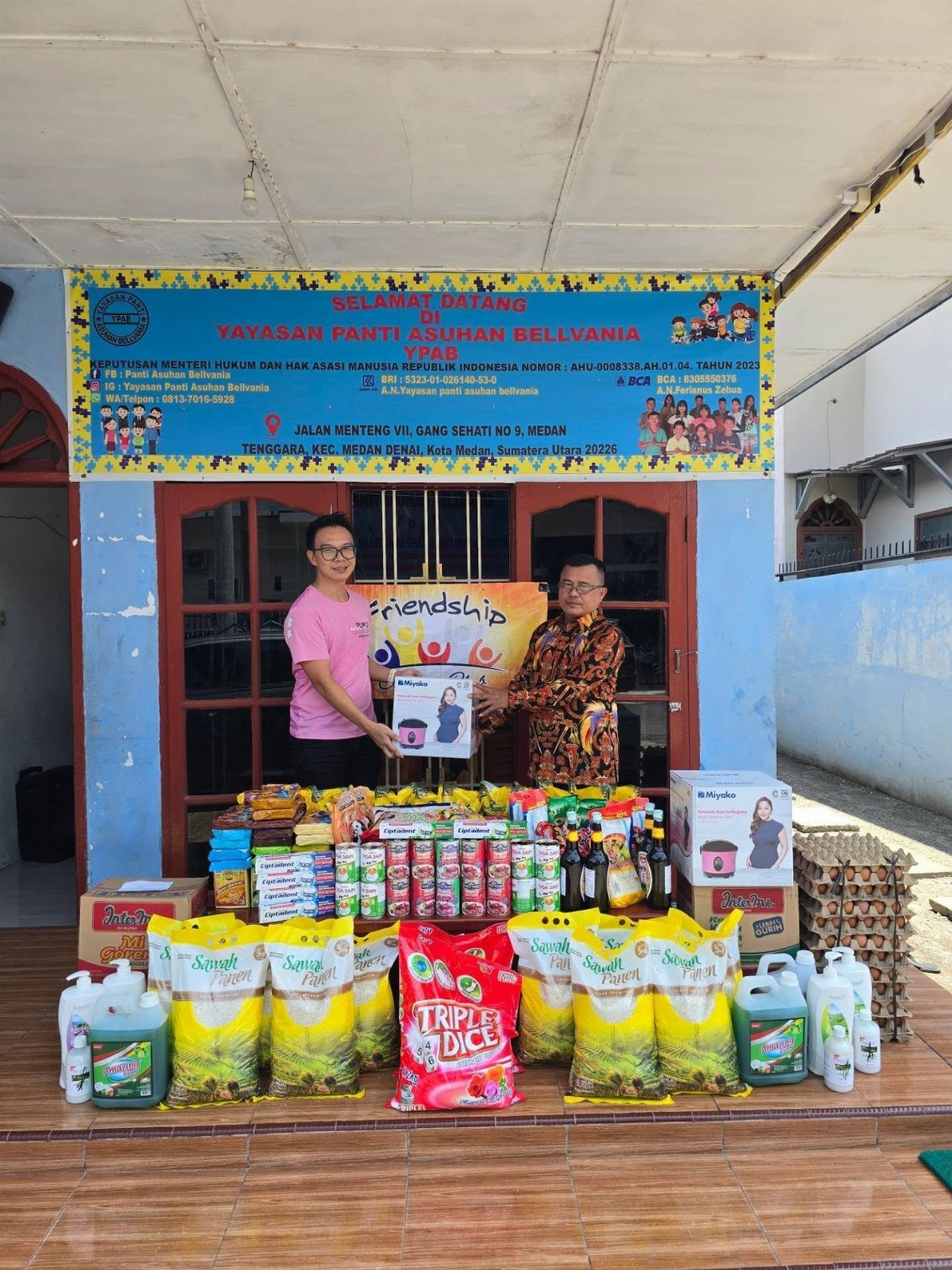 Two men posing behind large piles of donated food and supplies outdoors.