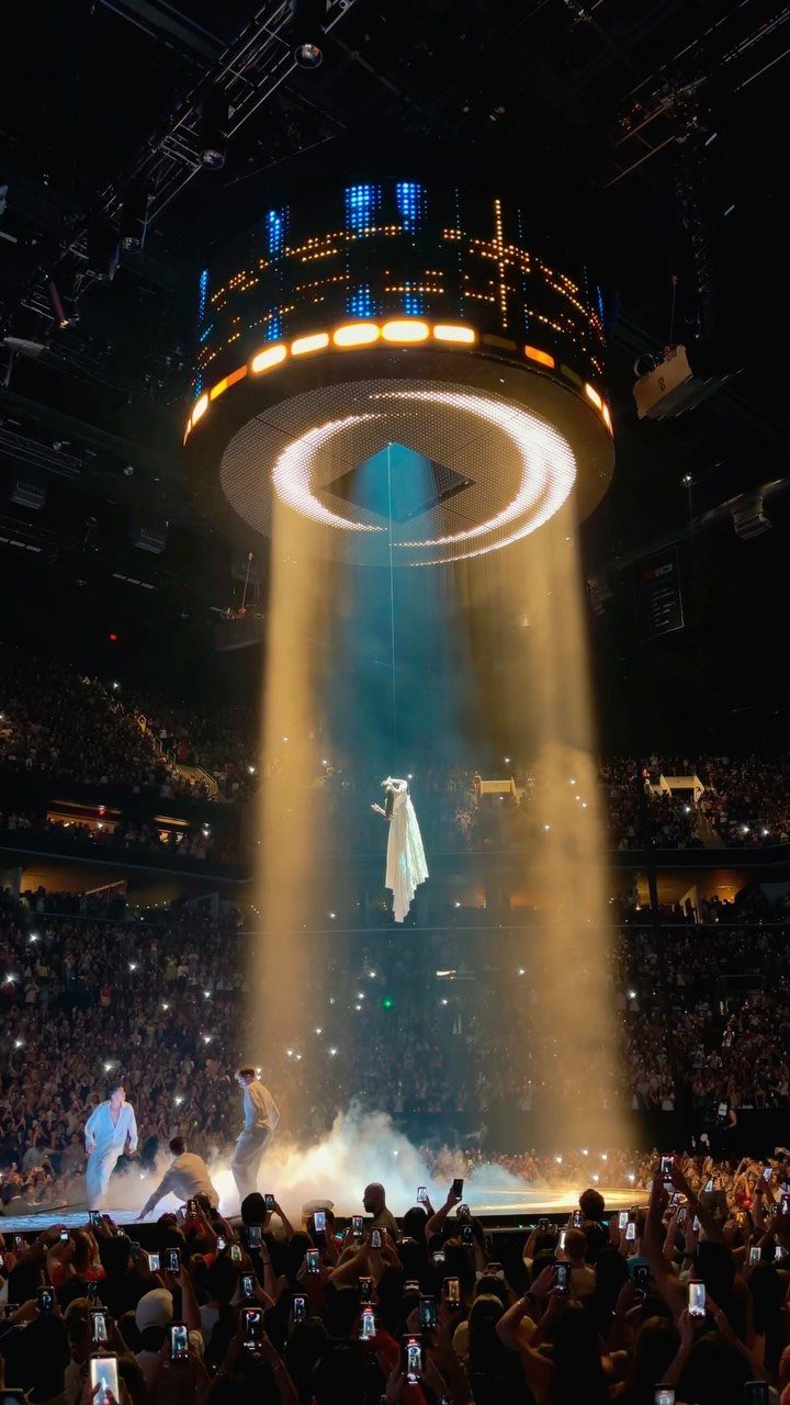 A performer stands on a smoky stage under bright spotlights in a crowded concert arena.