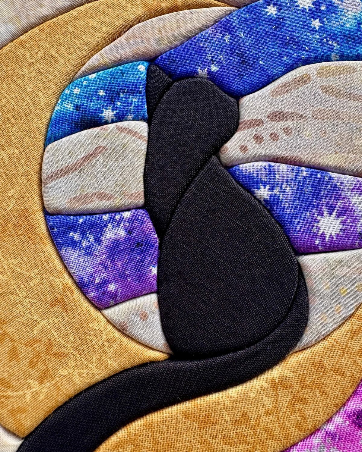 Fabric applique artwork showing a black cat silhouette on colorful patterned cloth.