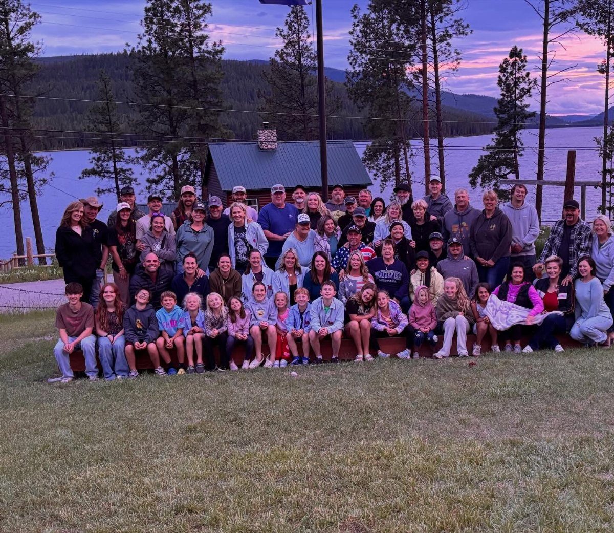 A large multi-generational family poses for a group photo outdoors near a lake.