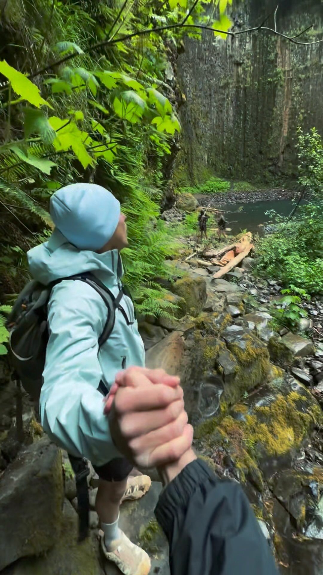 A hiker in a blue jacket looks up at a massive mossy cliffside while holding hands.