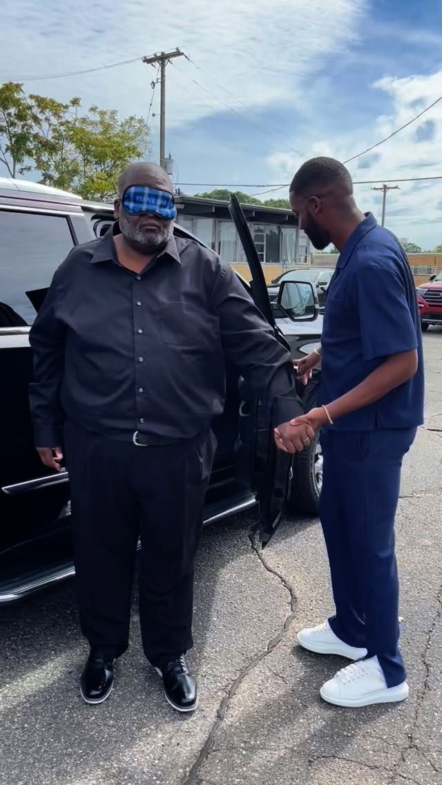 An older man wearing a sleep mask stands by a black car while being assisted.