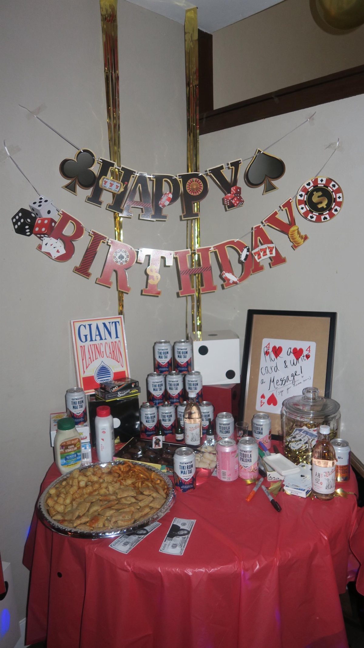 A red-clothed table decorated for a birthday with playing card themed banners, pastries, and assorted drinks.
