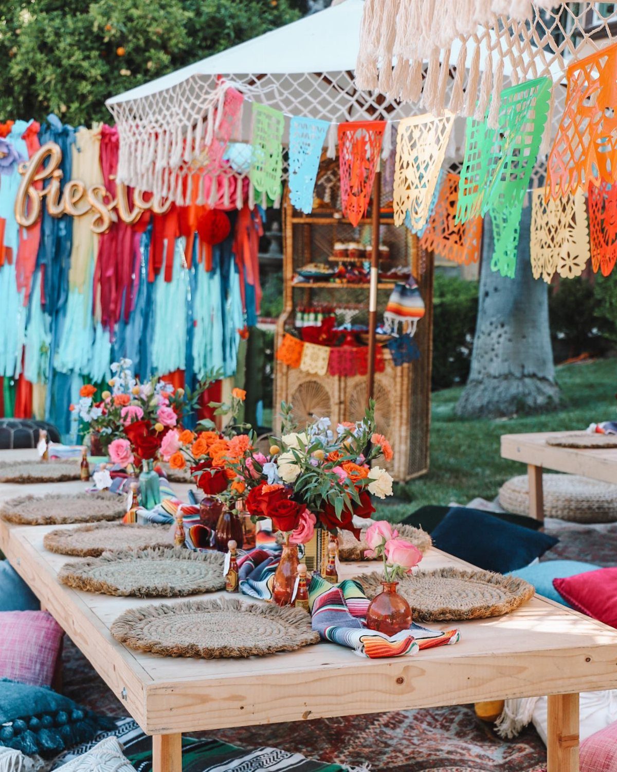 A festive outdoor low table setting with colorful flowers, woven placemats, and vibrant Mexican decorations.