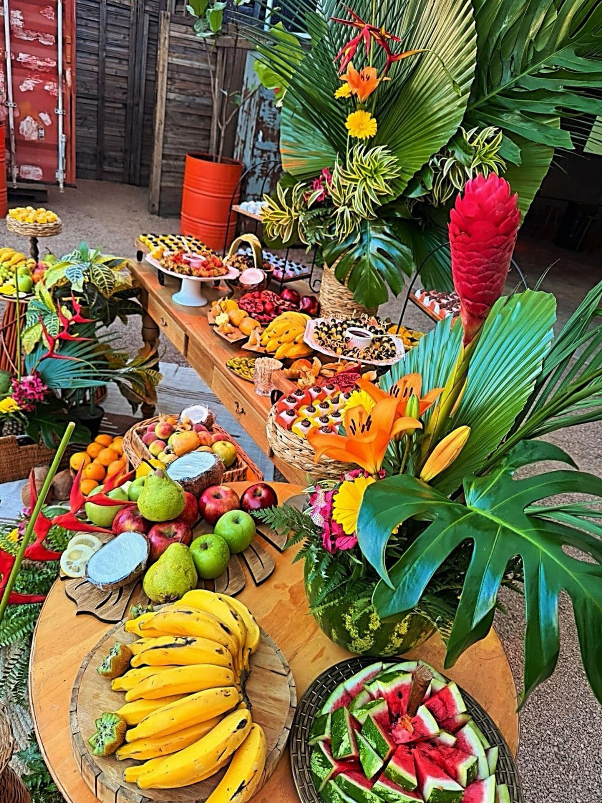 Tropical fruit platters and vibrant floral arrangements set on wooden tables for a luau theme.