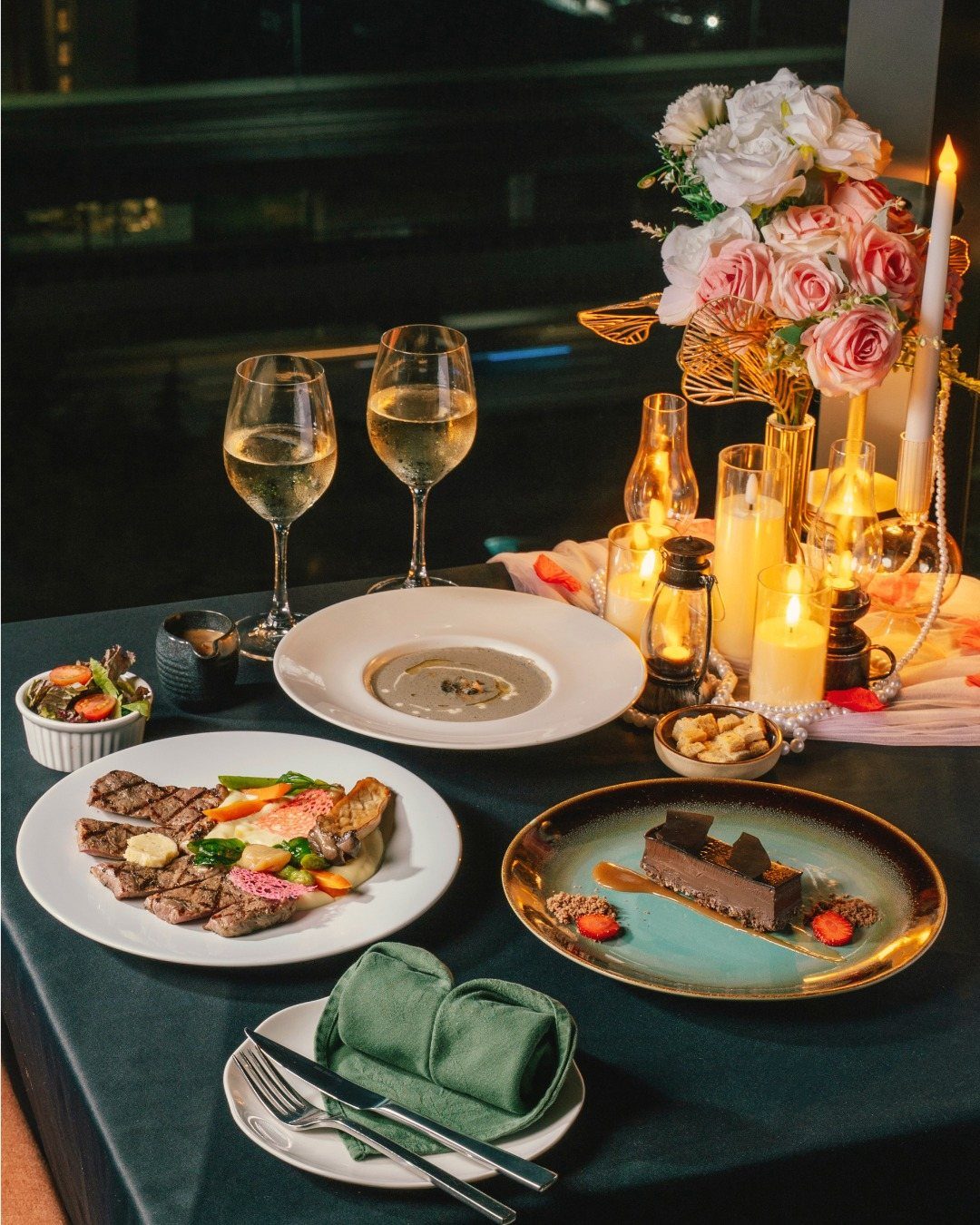 A formal dinner table set with steak, soup, dessert, wine, and lit candles.
