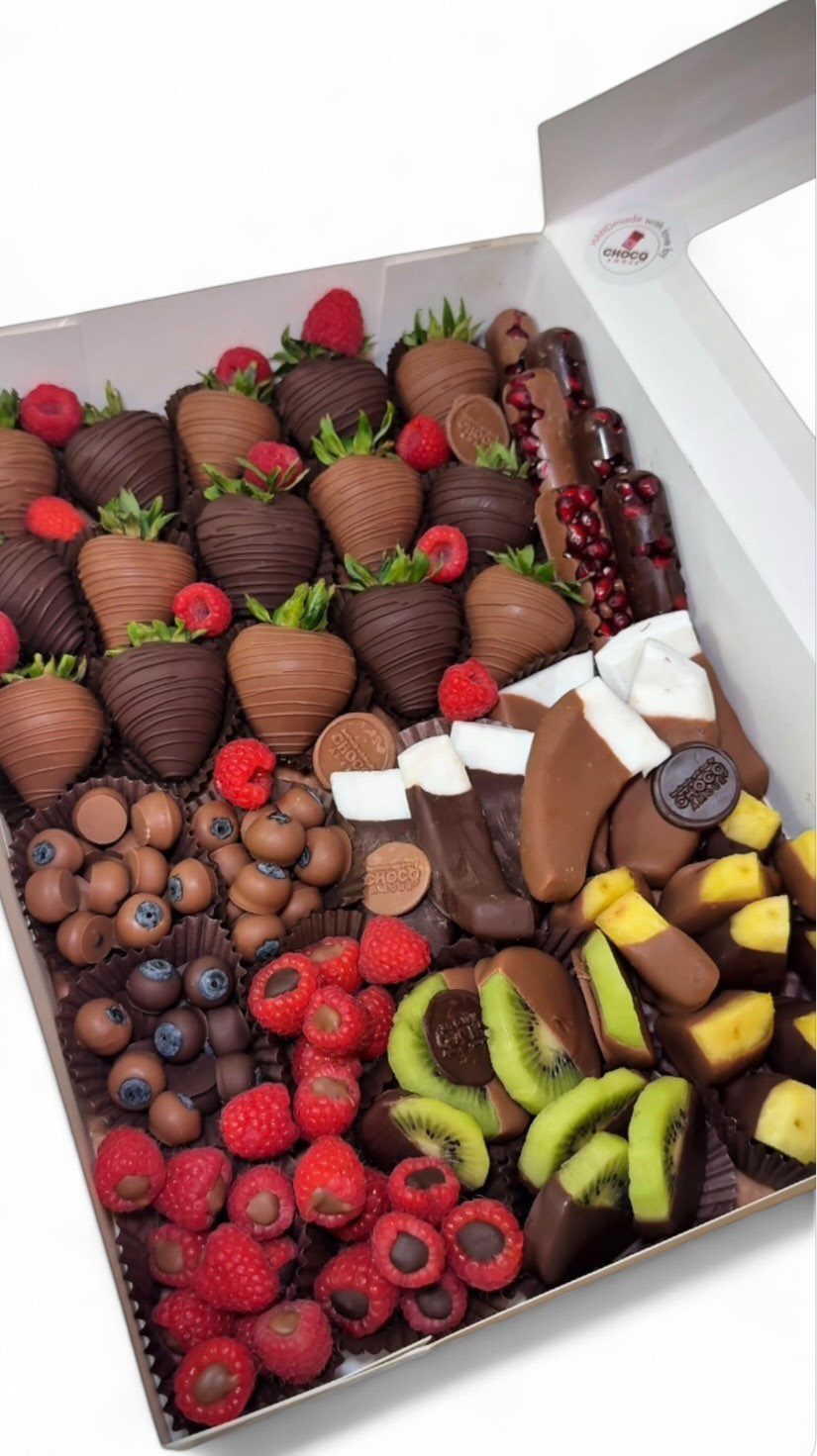 Chocolate-covered strawberries, kiwi, and bananas arranged in a white gift box with fresh berries.