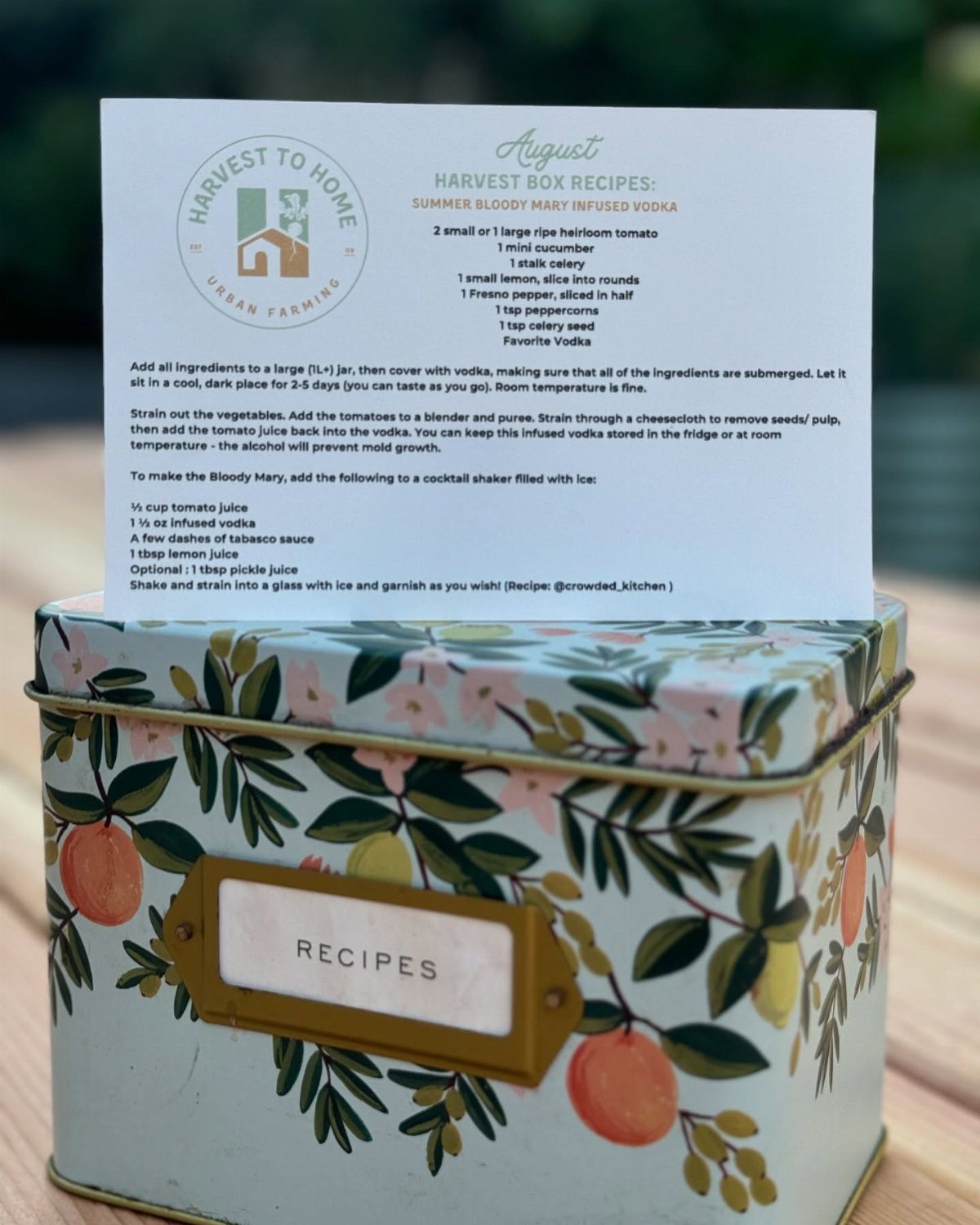 A printed recipe card for infused vodka resting on a botanical patterned metal recipe tin.