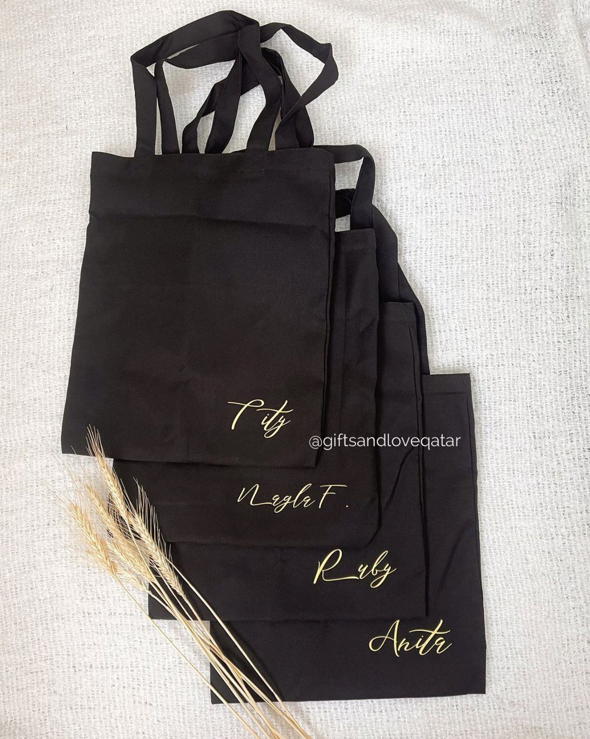 Four black personalized tote bags with gold script names resting on a white fabric background.