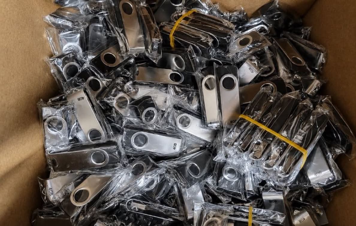 Many silver metal USB flash drives wrapped in clear plastic inside a cardboard box.