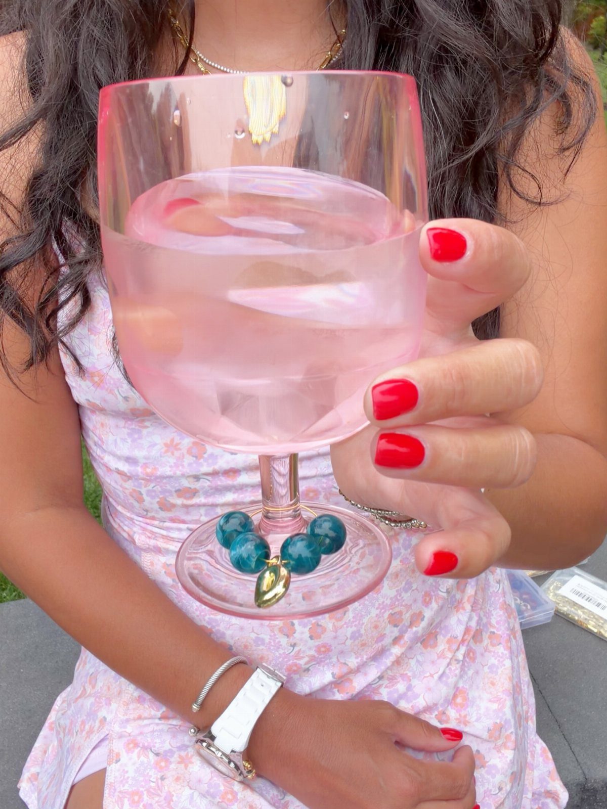 A hand with red nails holding a wine glass with a blue beaded charm.