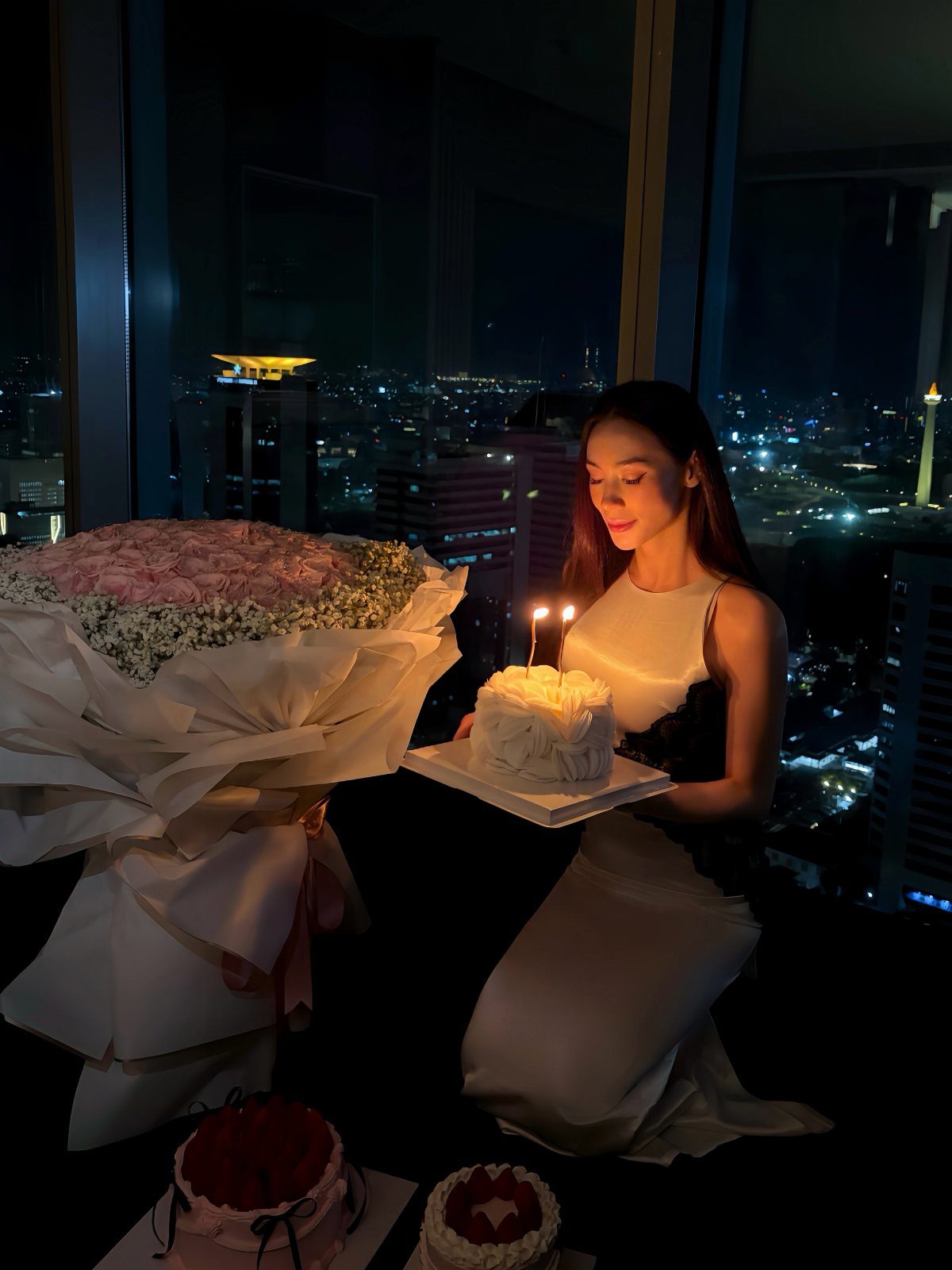 Woman in a white dress holding a small lit birthday cake against a city night view.