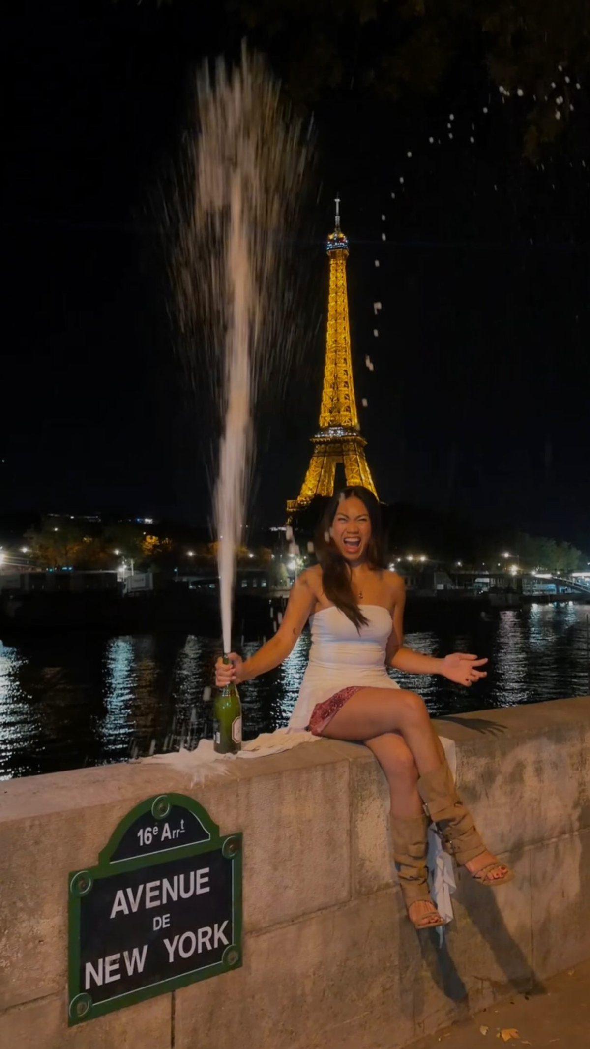 A woman laughs while popping a champagne bottle with a large spray in front of the Eiffel Tower.
