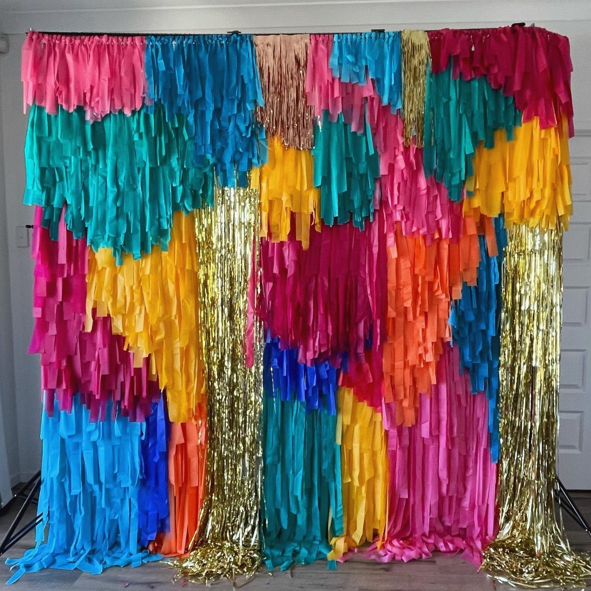 A vibrant multi-colored paper fringe backdrop with gold metallic tinsel strips hanging from a stand.