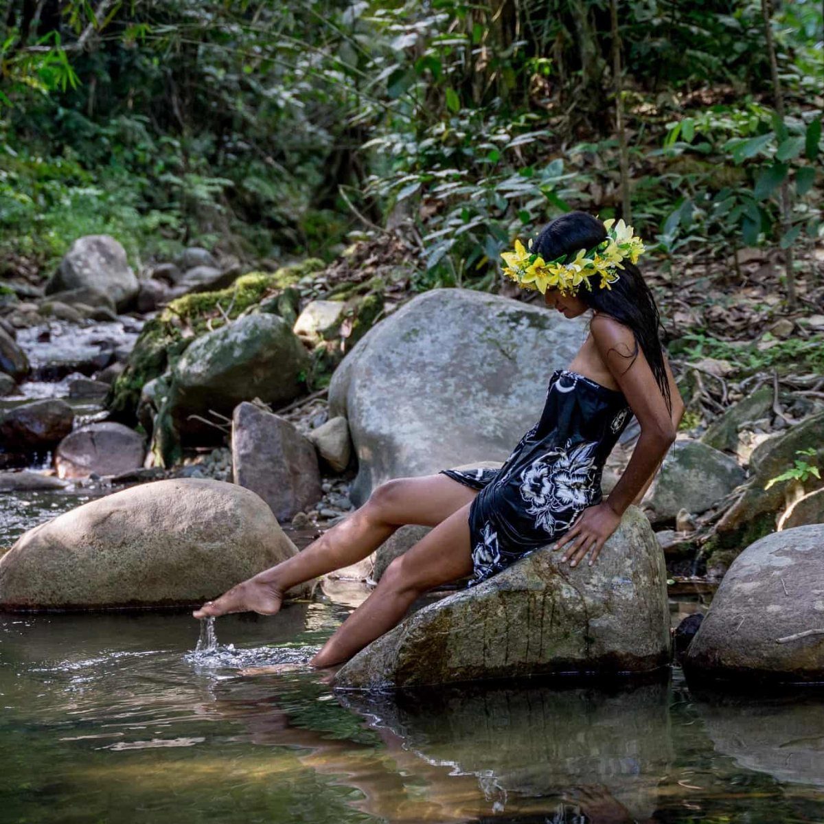 A woman wearing a flower crown sits on a rock by a forest stream.