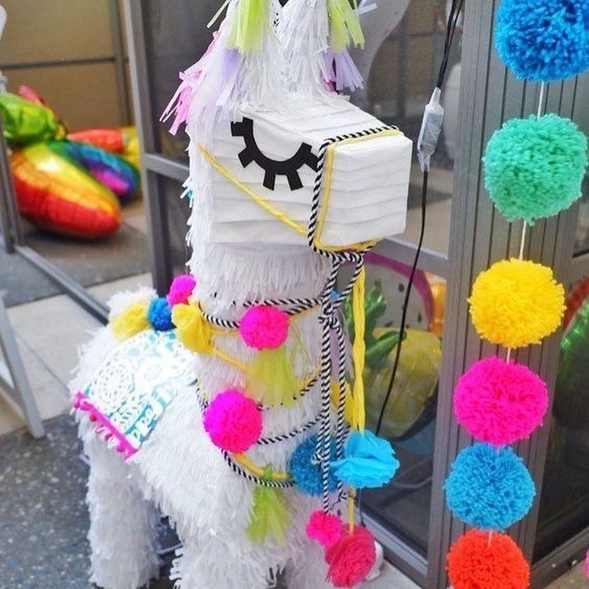 A white llama piñata decorated with bright, colorful pom-poms standing outdoors.