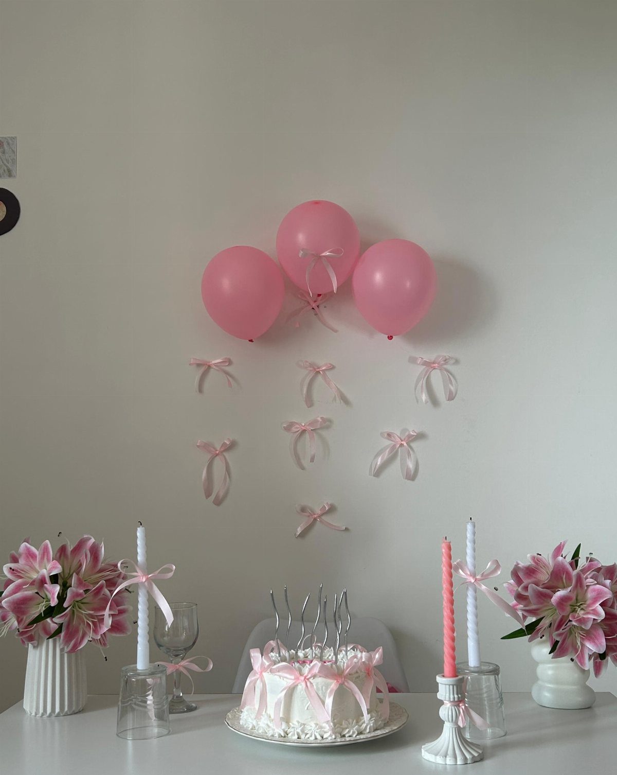 Pink balloons, white cake with bows, and pink lilies arranged on a white table.