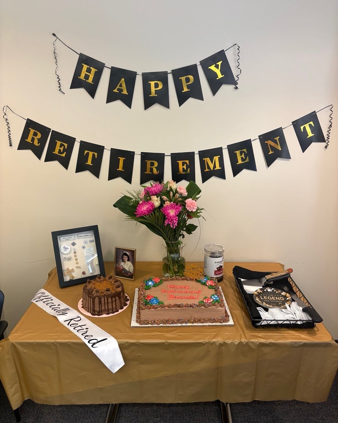 A retirement celebration table with two chocolate cakes, flowers, and framed photos under retirement banners.