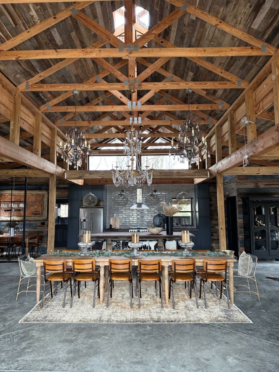A long wooden dining table with chairs set inside a large rustic barn with timber ceilings.