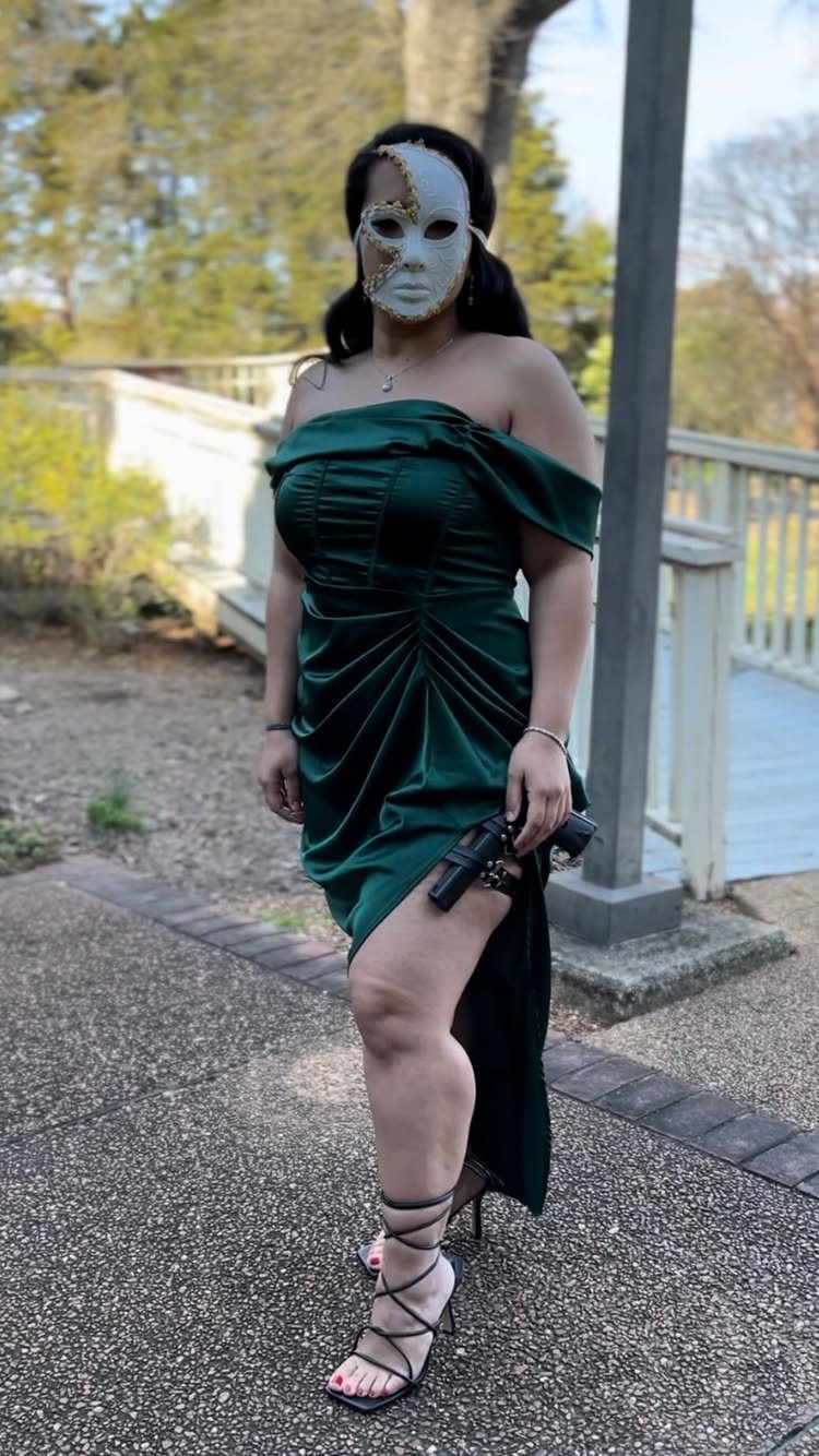 Woman in a green dress and white mask holding a toy gun outdoors.