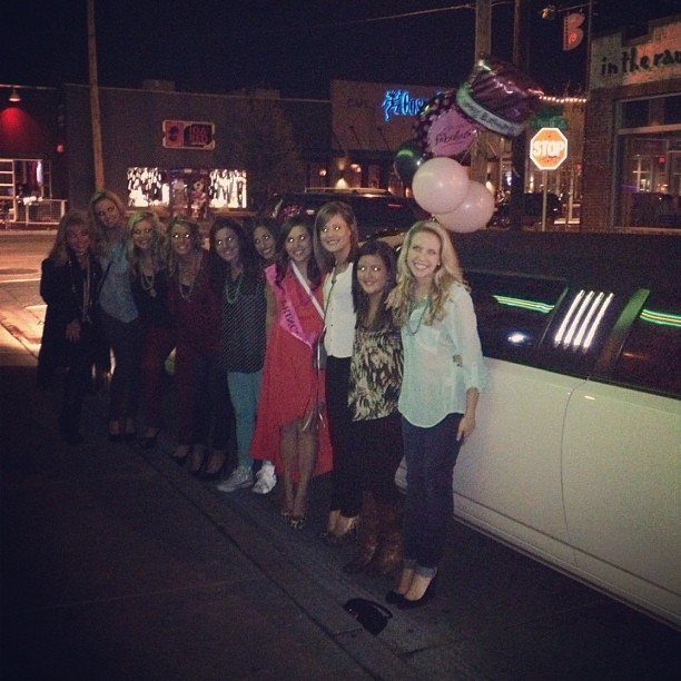 A group of smiling young women posing next to a white limousine at night.