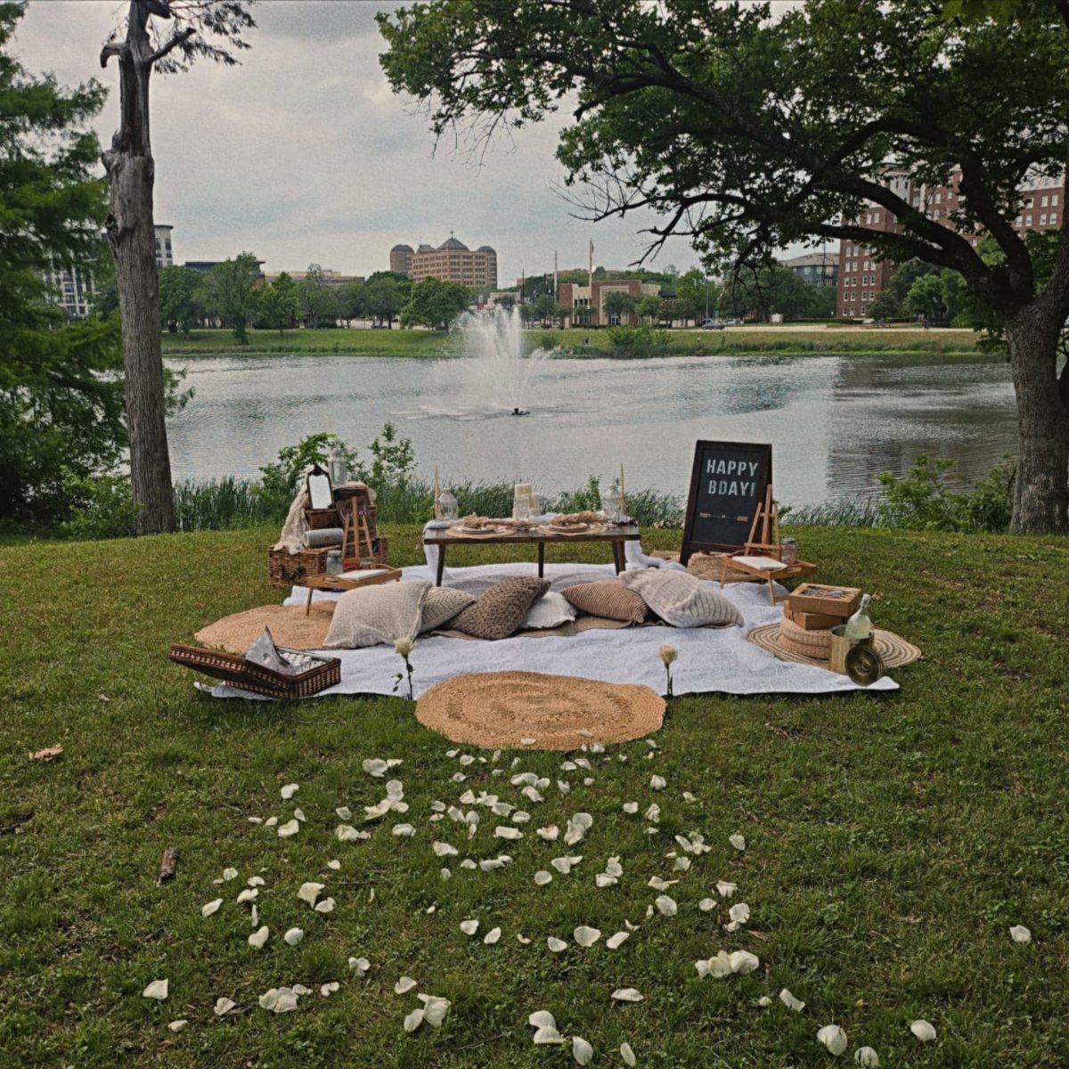 A decorated picnic setup with pillows, a small table, and flower petals by a lake.