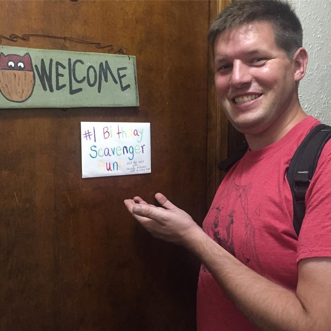 A smiling man in a red shirt gestures toward a birthday scavenger hunt sign on a wooden door.
