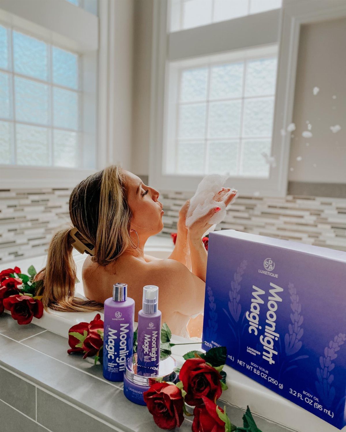 Woman blowing soap bubbles in a bathtub surrounded by red roses and purple beauty products.